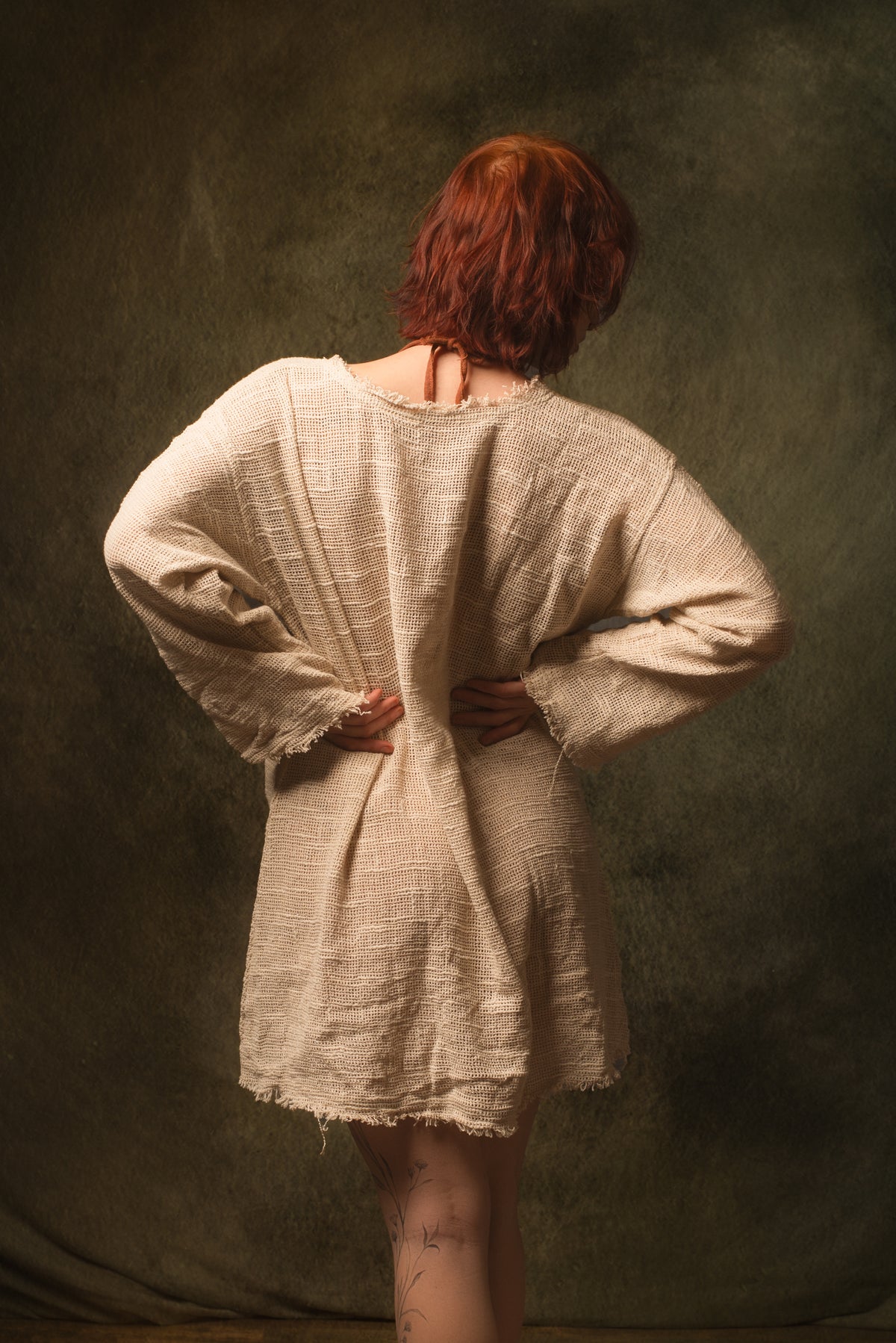 A person with short red hair stands with their back to the camera wearing a loose, textured beige linen tunic dress with long sleeves and raw, frayed edges, posed against a dark, textured studio backdrop.