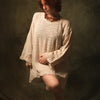 A model with short red hair wearing a loose, textured cream-colored linen tunic dress, posing against a dark, textured studio backdrop.