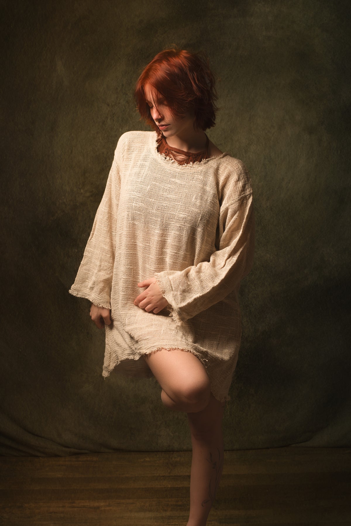A model with short red hair wearing a loose, textured cream-colored linen tunic dress, posing against a dark, textured studio backdrop.
