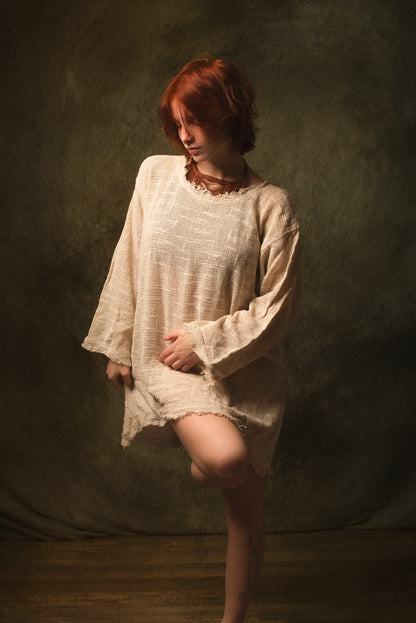 A model with short red hair wearing a loose, textured cream-colored linen tunic dress, posing against a dark, textured studio backdrop.
