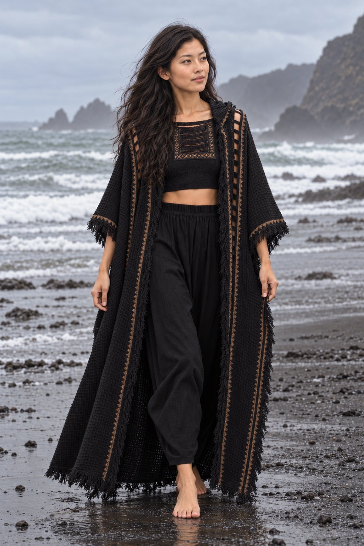 A woman stands barefoot on a dark volcanic beach wearing a black textured bohemian-style outfit consisting of a long open-front cardigan with gold trim, a black crop top, and black wide-leg pants, with ocean waves and rocky cliffs in the background.