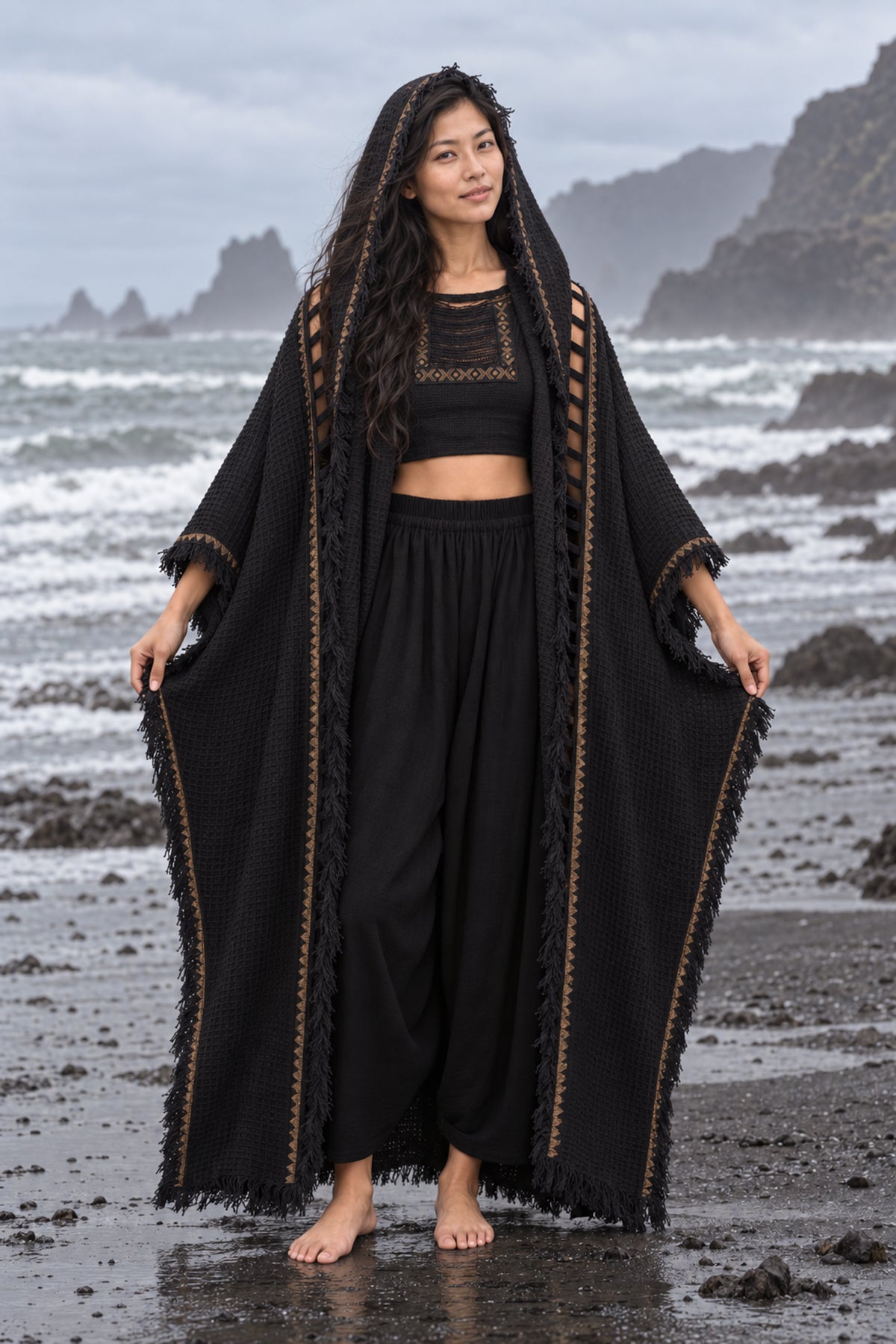 A woman stands barefoot on a rocky beach wearing a flowing black outfit with a long open-front draped outer layer, black cropped top, and black wide-leg pants. The background shows ocean waves, dark rocks, and a cloudy sky.