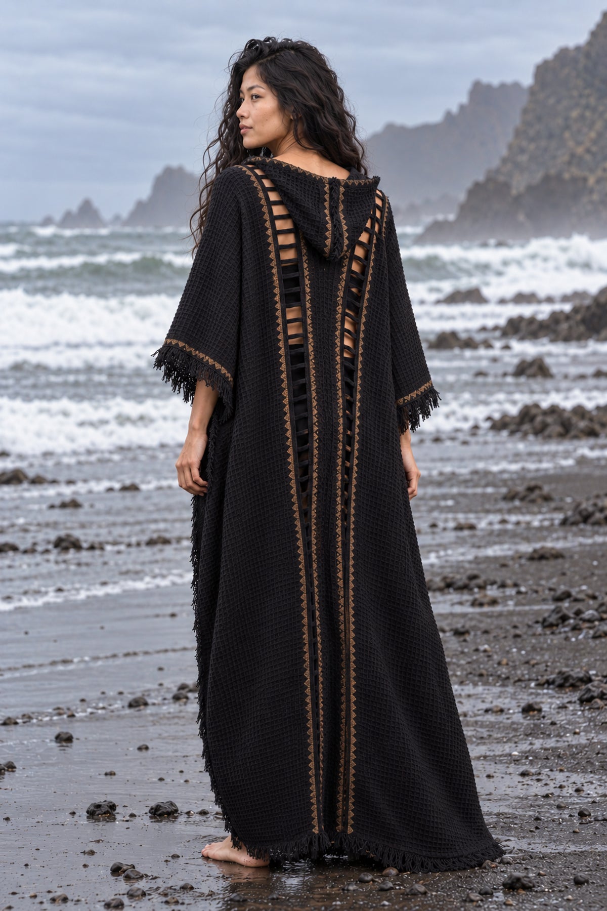 A woman stands on a rocky beach wearing a long black textured kaftan dress with gold trim and side cutouts, photographed from the back against an ocean backdrop.