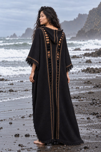 A woman stands on a rocky beach wearing a long black textured kaftan dress with gold trim and side cutouts, photographed from the back against an ocean backdrop.