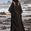 A woman stands barefoot on a rocky beach facing the ocean, wearing a long black hooded outer garment with cutout side panels and gold-toned vertical trim.