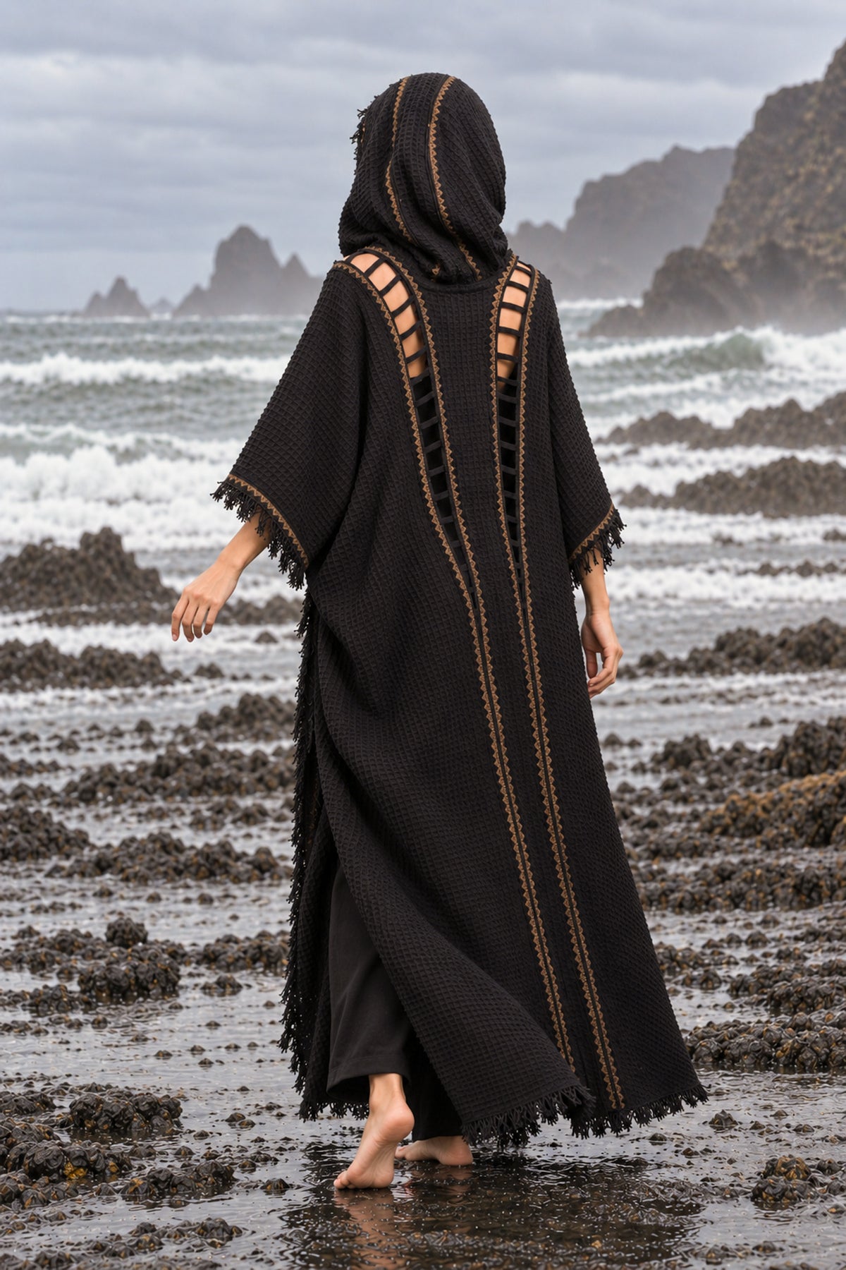 A woman stands barefoot on a rocky beach facing the ocean, wearing a long black hooded outer garment with cutout side panels and gold-toned vertical trim.