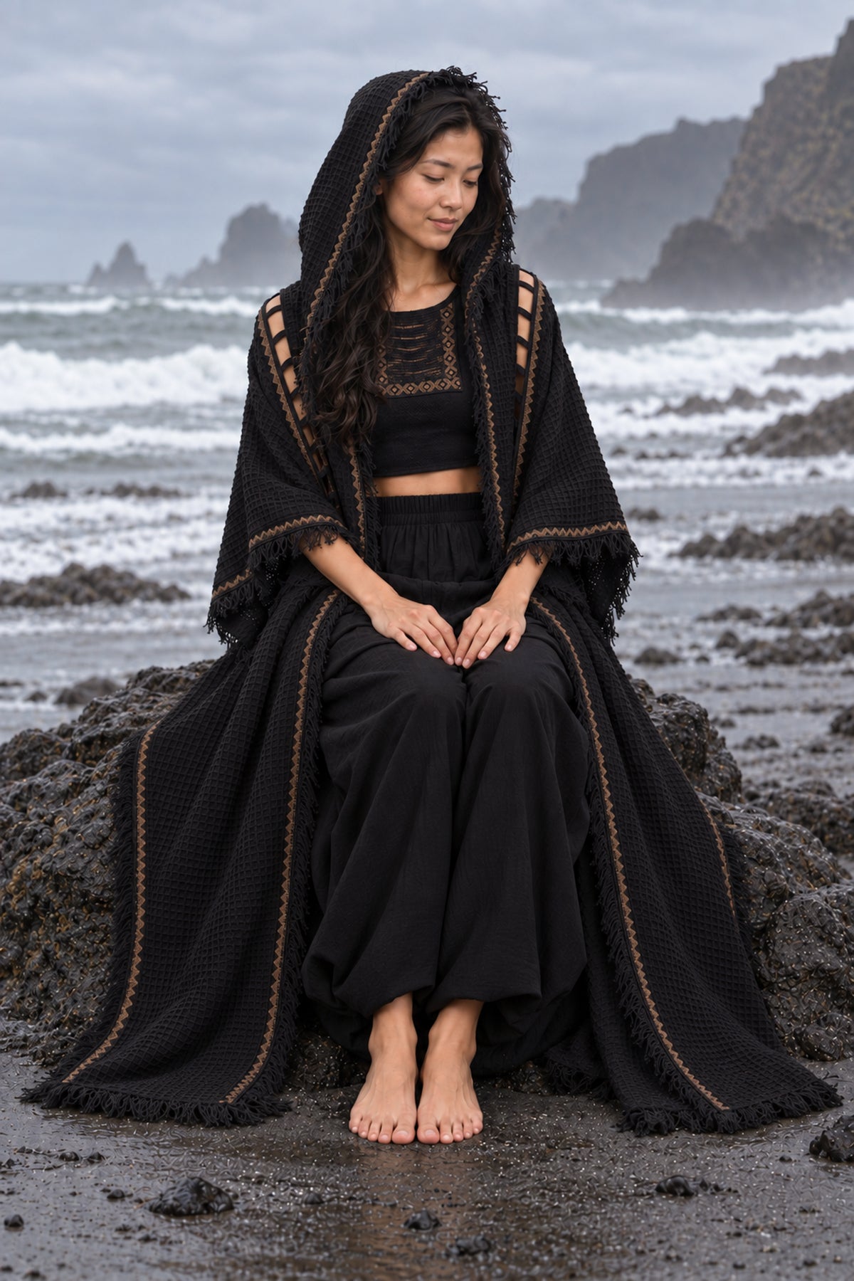 A woman sits barefoot on a dark volcanic beach wearing a black textured waffle-knit hooded poncho with gold geometric embroidery over a matching black two-piece outfit.