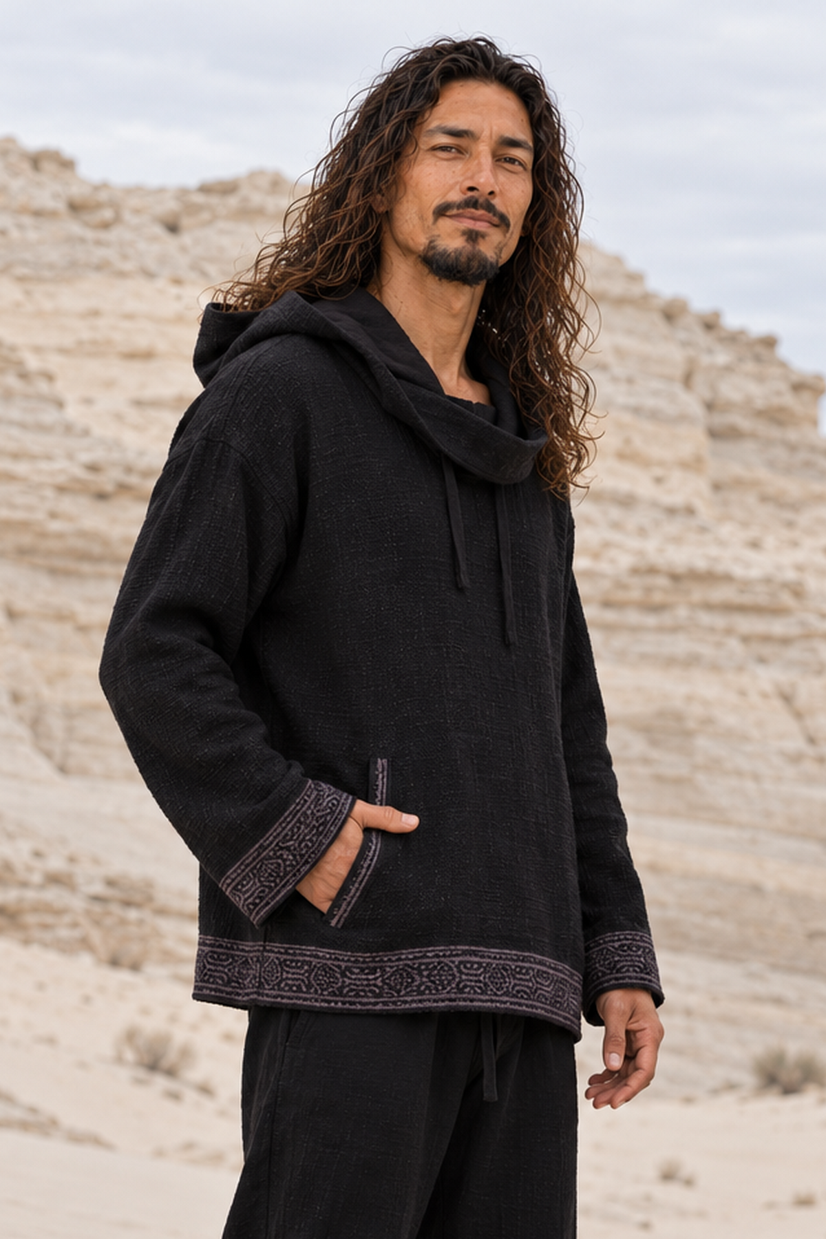 Man in black handwoven cotton hoodie and black pants, one hand in pocket, hood down, in a sandstone canyon