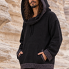 Man in black SUKKUN handwoven cotton hoodie, hood up and hands in pockets, grey block-printed trim, by sandstone cliffs