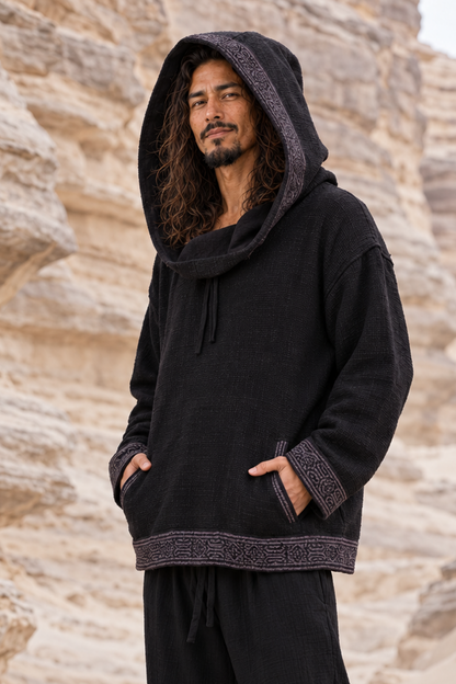Man in black SUKKUN handwoven cotton hoodie, hood up and hands in pockets, grey block-printed trim, by sandstone cliffs