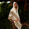 A man wearing a white wool poncho with tribal embroidery, hood up, standing outdoors.